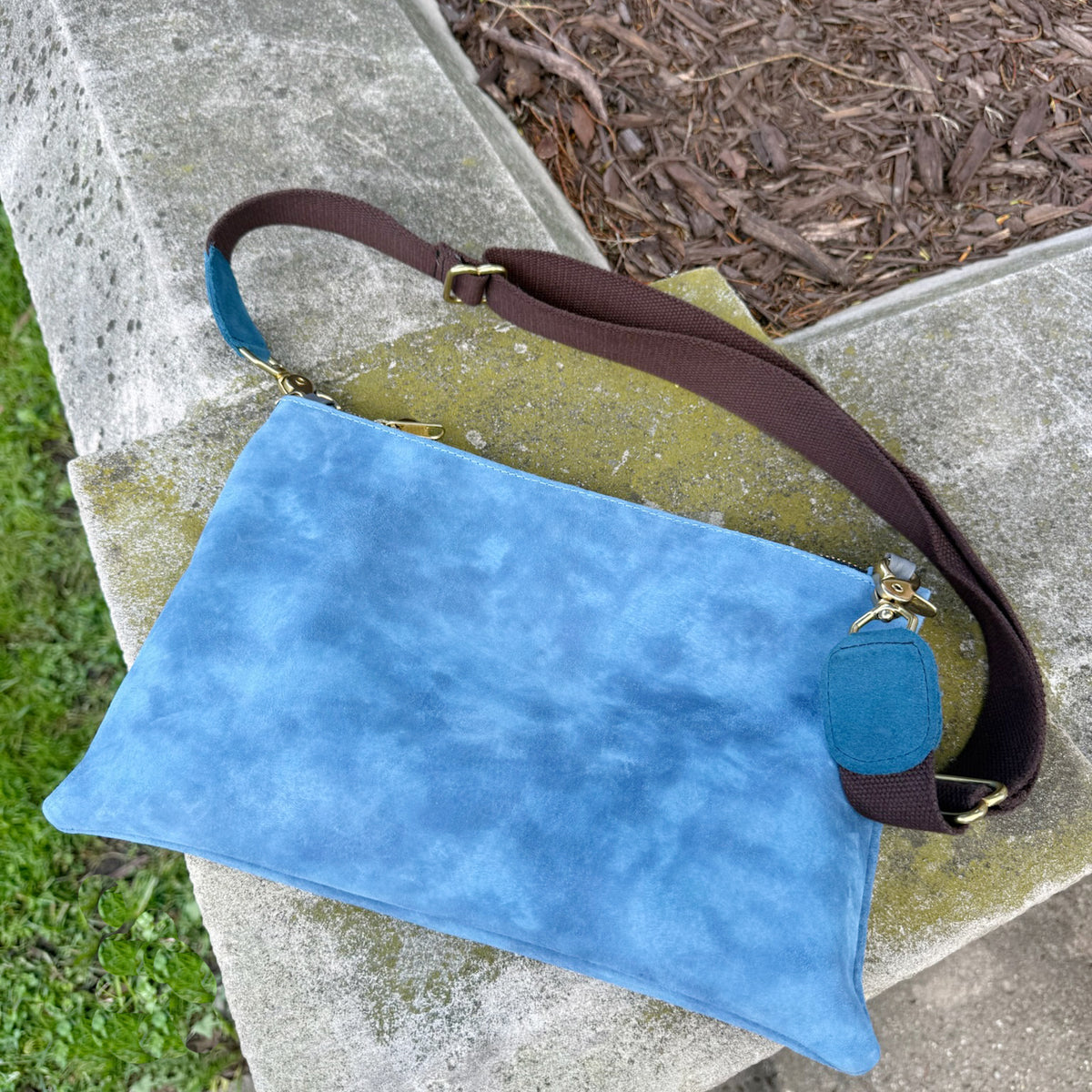 A blue leather and suede clutch bag with a brown shoulder strap and gold-tone hardware, resting on a concrete garden bench with mulch and grass visible in the background.