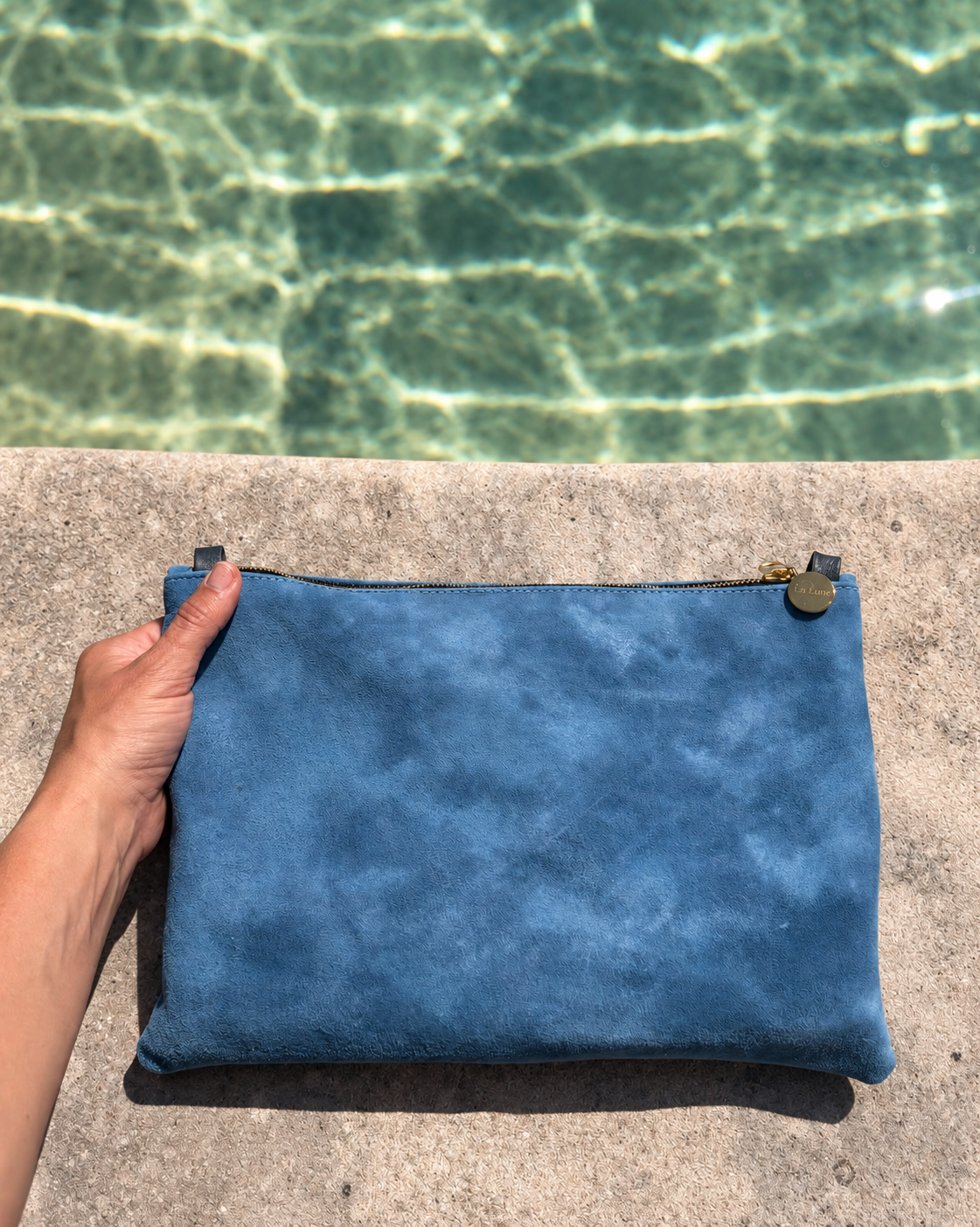 A person's hand holds a blue suede zippered pouch against a concrete ledge with a swimming pool background visible above.